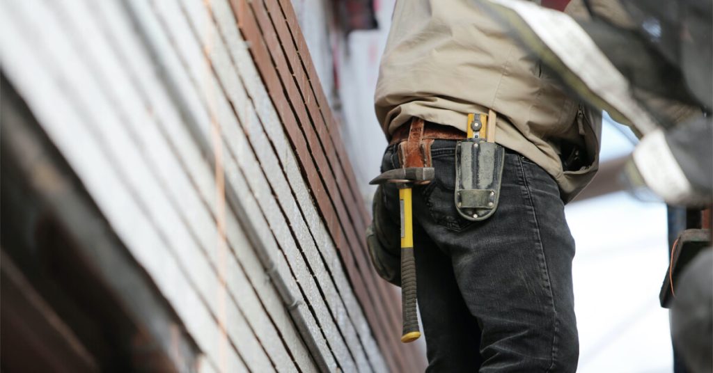 trabalhador da área de profissionais de construção civil de calça escura e camiseta cinza com um martelo pendurado ao cinto